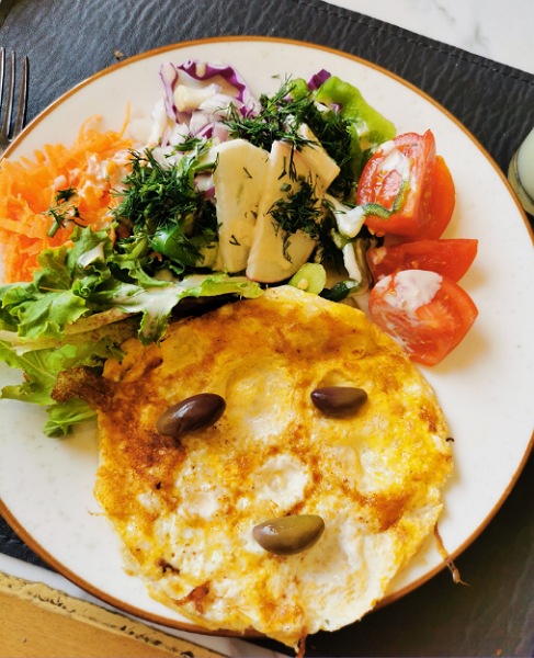 Frühstück Omlet als Gesicht, Oliven sind Augen und Mund und Salat, Tomaten, geriebene Karotten die Haare.